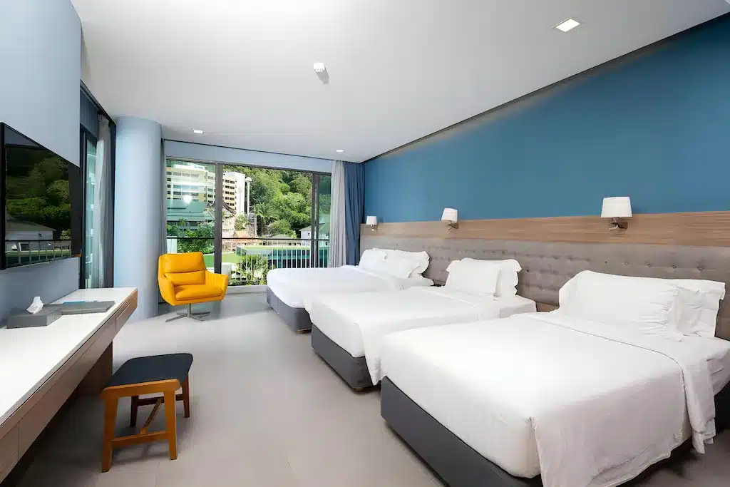 ห้องพักใน ที่พักกระบี่ โรงแรมทันสมัยมีเตียง 3 เตียงที่จัดอย่างประณีต โต๊ะทำงาน เก้าอี้เท้าแขนสีเหลืองข้างหน้าต่าง ผนังสีฟ้า และทิวทัศน์ของต้นไม้และอาคารต่างๆ เหมาะสำหรับผู้ที่ต้องการ โรงแรมกระบี่ ในบรรยากาศที่ผ่อนคลาย.