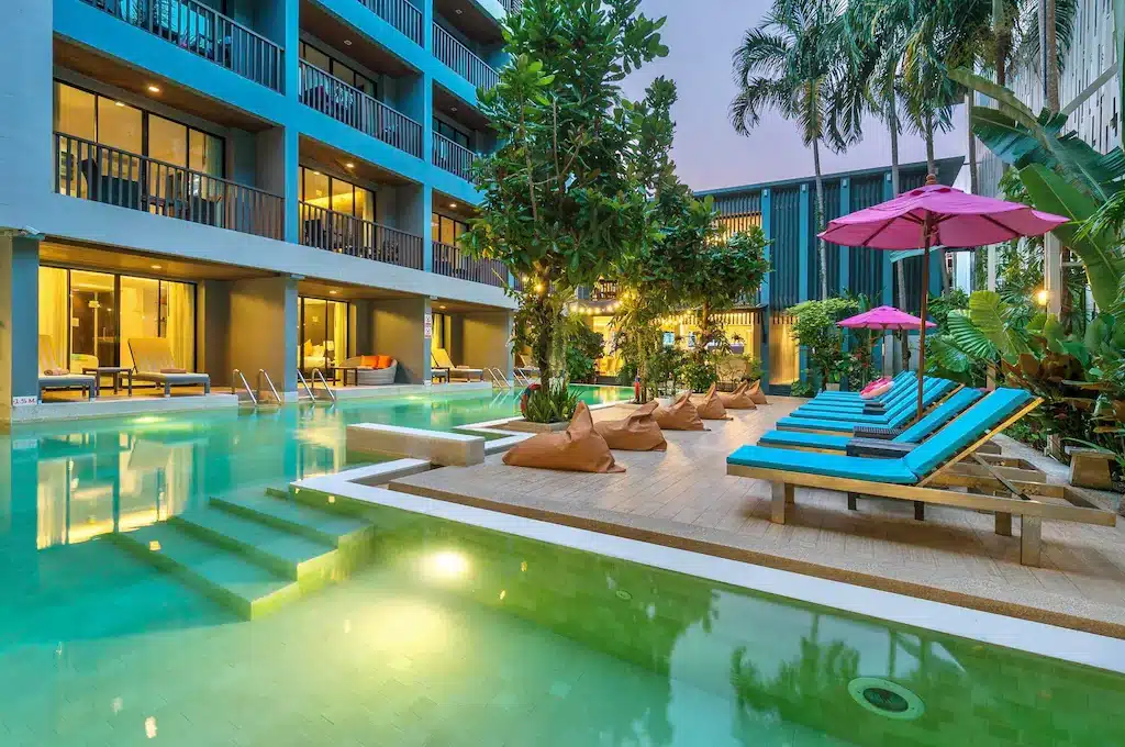 บริเวณสระว่ายน้ำของโรงแรมทันสมัยพร้อมเก้าอี้เลานจ์สีน้ำเงิน เก้าอี้บีน ที่พักอ่าวนางกระบี่ แบ็กสีน้ำตาล และร่มสีชมพู รายล้อมด้วยต้นไม้สีเขียวและอาคารหลายชั้น เหมาะสำหรับการพักผ่อนที่รีสอร์ทสุดเก๋ไก๋ในกระบี่.