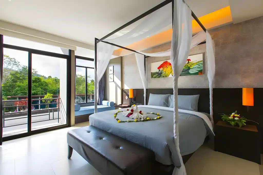 ห้องพักในโรงแรมทันสมัยพร้อมเตียงกระโจม ชุดเครื่องนอนสีเทา ผ้าเช็ดตัว ทางเข้าระเบียง หน้าต่างบานใหญ่ และต้นไม้เขียวขจีด้านนอก เหมาะสำหรับผู้ที่กำลังมองหาประสบการณ์ที่พักสไตล์รีสอร์ทกระบี่หรือกระบี่.
