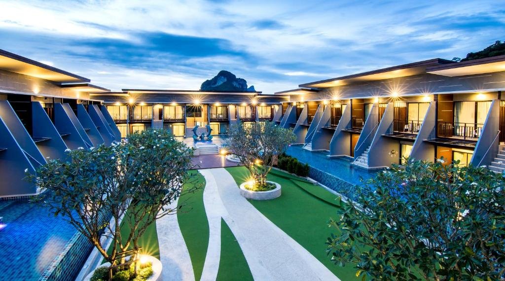 ลานภายในโรงแรม ที่พักกระบี่ ที่ทันสมัยพร้อมสถาปัตยกรรมแบบสมมาตร สวนภูมิทัศน์ และสระว่ายน้ำในยามพลบค่ำ ส่องสว่างด้วยไฟกลางแจ้ง พร้อมภูเขาที่มองเห็นได้เป็นฉากหลัง โรงแรมกระบี่ ที่เหมาะสำหรับการเข้าพักของคุณ.