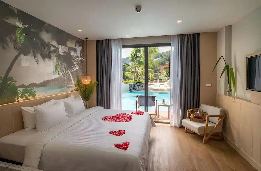 ห้องพักในโรงแรม ที่พักกระบี่ ที่มีเตียงขนาดใหญ่ตกแต่งด้วยกลีบดอกไม้สีแดง ภาพฝาผนังชายหาด พื้นที่นั่งเล่น ทีวี และประตูกระจกที่เปิดออกไปเห็นวิวสระว่ายน้ำ เหมาะสำหรับการเข้าพักของคุณในรีสอร์ทกระบี่หรือใครก็ตามที่กำลังมองหาที่พักที่มีสไตล์กระบี่.