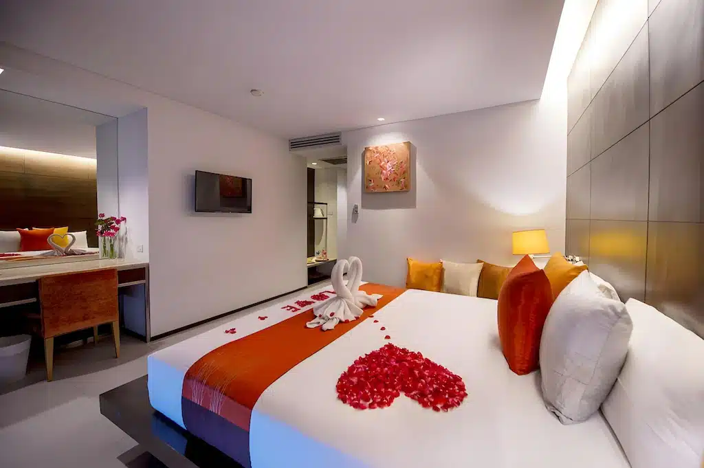 ห้องพักในโรงแรมทันสมัยพร้อมเตียงคิงไซส์ ตกแต่งด้วยสีส้ม ผ้าเช็ดตัวรูป ที่พักอ่าวนางกระบี่ หงส์ กลีบกุหลาบรูปหัวใจ โต๊ะทำงานพร้อมดอกไม้ งานศิลปะบนผนัง และแสงไฟอันอบอุ่น เหมาะสำหรับการเข้าพักของคุณที่อ่าวนางกระบี่.