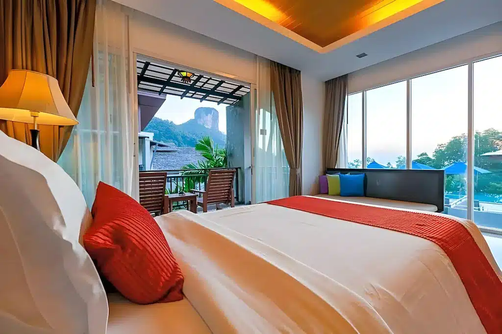 ห้องพักในโรงแรมทันสมัยพร้อมเตียงนอนที่จัดอย่างประณีต ตกแต่งด้วยสี ที่พักกระบี่ แดง และระเบียงเล็กๆ ที่มองเห็นภูเขาและต้นไม้เขียวขจี เหมาะสำหรับผู้ที่กำลังมองหาที่พักที่ผ่อนคลาย พร้อมวิวทิวทัศน์ที่สวยงามและความสบาย.