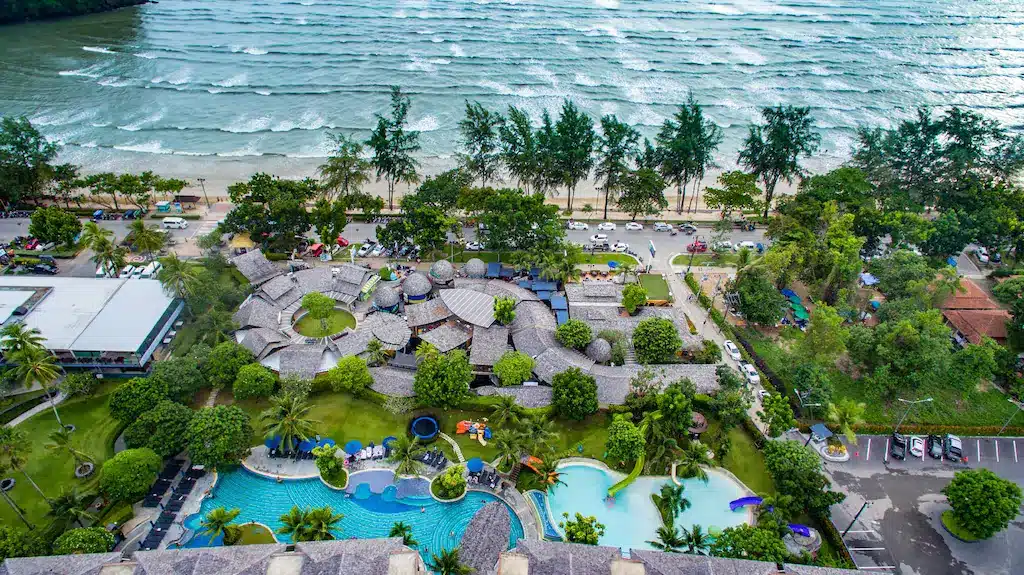 มุมมองทางอากาศของรีสอร์ทริม ที่พักอ่าวนางราคาถูก ชายหาดที่อ่าวนางกระบี่ มีทั้งสระน้ำคดเคี้ยว อาคารทรงกลม ต้นปาล์ม และแนวชายฝั่งทะเลคลื่นที่ล้อมรอบด้วยถนนที่มีต้นไม้เรียงรายและรถยนต์ที่จอดอยู่.