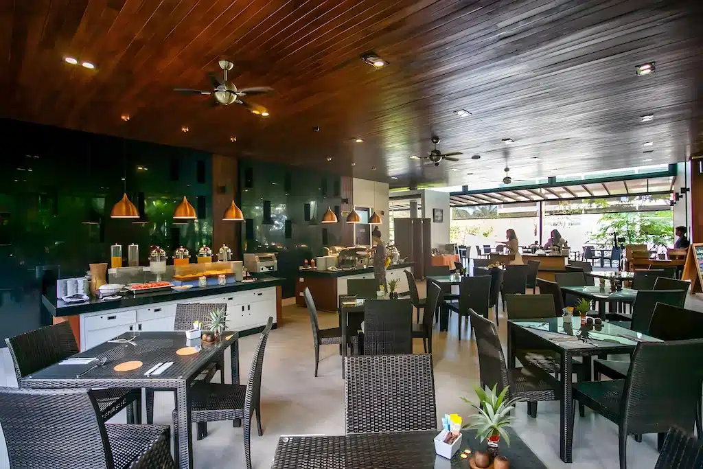 การตกแต่งภาย โรงแรมกระบี่ ในร้านอาหารสไตล์โมเดิร์นที่รีสอร์ทกระบี่ประกอบด้วยเก้าอี้หวายสีดำ โต๊ะกระจก อุปกรณ์บุฟเฟ่ต์ เพดานไม้ โคมไฟระย้า และหน้าต่างบานใหญ่ที่เปิดรับแสงธรรมชาติ.