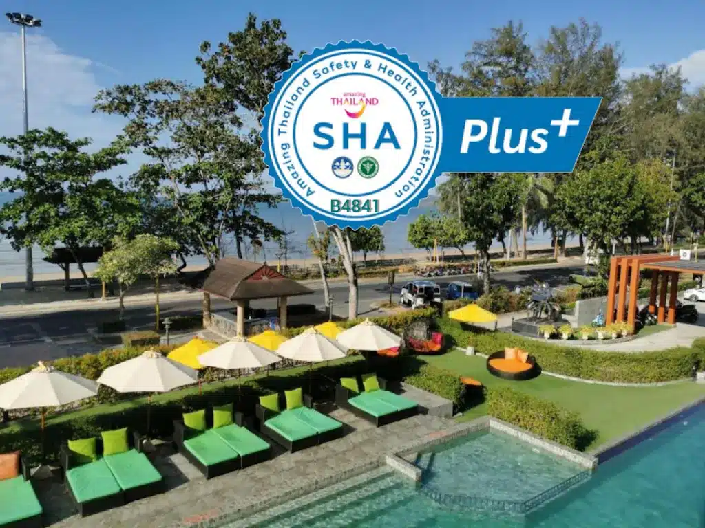 บริเวณสระว่ายน้ำของรีสอร์ทที่ รีสอร์ทกระบี่ มีเก้าอี้เลานจ์สีเขียว ร่มสีเบจ และต้นไม้ใกล้ชายหาด พร้อมด้วยป้ายรับรองความปลอดภัยและสุขภาพ "SHA Plus+" เพื่อความอุ่นใจของคุณ.