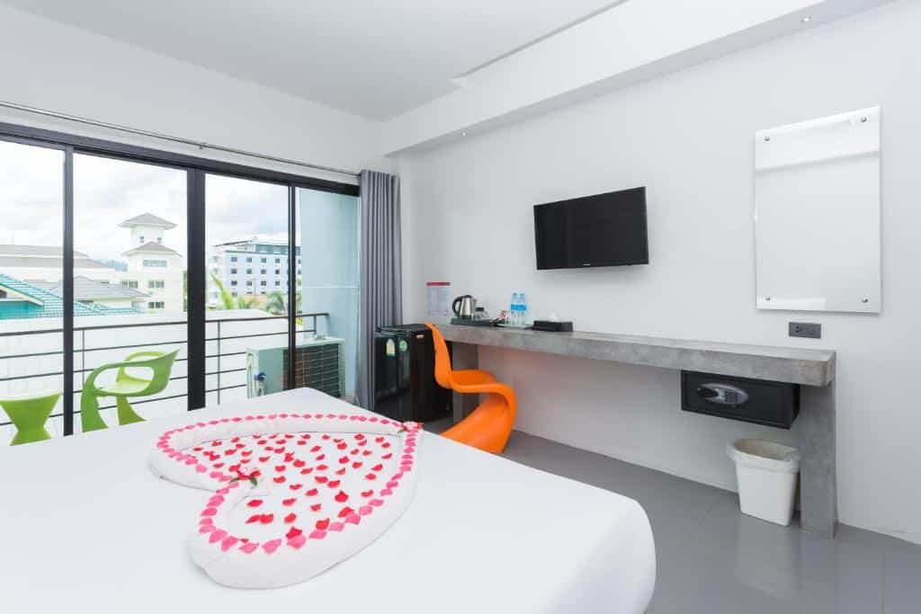 ห้องพักโรงแรม ที่พักกระบี่ ทันสมัยที่โรงแรมกระบี่ มีเตียงนอนที่ประดับด้วยผ้าขนหนูลายหงส์และกลีบกุหลาบรูปหัวใจ โต๊ะทำงานพร้อมเก้าอี้สีส้ม ทีวี ตู้เย็นขนาดเล็ก และระเบียงพร้อมที่นั่งกลางแจ้งสำหรับการพักผ่อนที่ผ่อนคลาย.
