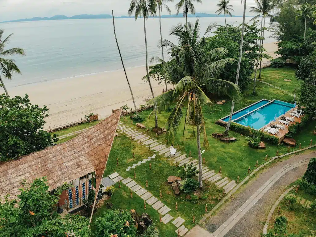 วิวทางอากาศของรีสอร์ทกระบี่ริม ที่พักกระบี่ ชายหาดพร้อมอาคารหลังคามุงจาก สระว่ายน้ำ ต้นปาล์ม และหาดทรายริมน้ำทะเลอันเงียบสงบ.