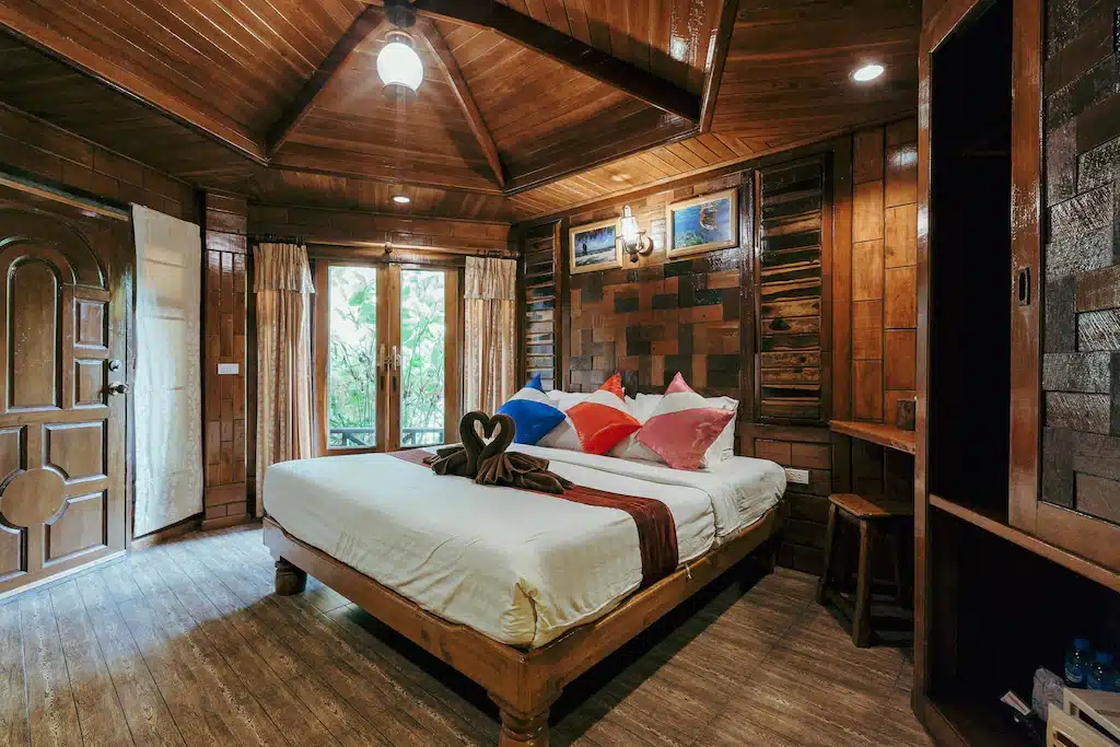 ห้องนอนไม้แสนสบายที่ พักอ่าวนาง พร้อมเตียงขนาดใหญ่ หมอนสีสัน ที่พักอ่าวนาง สดใส ผ้าเช็ดตัวพับเป็นรูปหัวใจ โต๊ะเขียนหนังสือ และแสงธรรมชาติที่ส่องเข้ามาทางหน้าต่างและประตูที่เปิดอยู่.