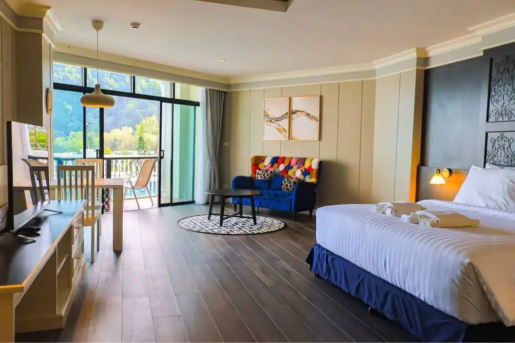 ห้องพัก โรงแรมกระบี่ โรงแรมเมืองกระบี่ที่กว้างขวางพร้อมเตียงขนาดใหญ่ โซฟาสีสันสดใส พื้นที่รับประทานอาหารเล็กๆ ทีวี และประตูกระจกที่เปิดออกสู่ระเบียงพร้อมทิวทัศน์อันเขียวขจีอันเงียบสงบภายนอก.