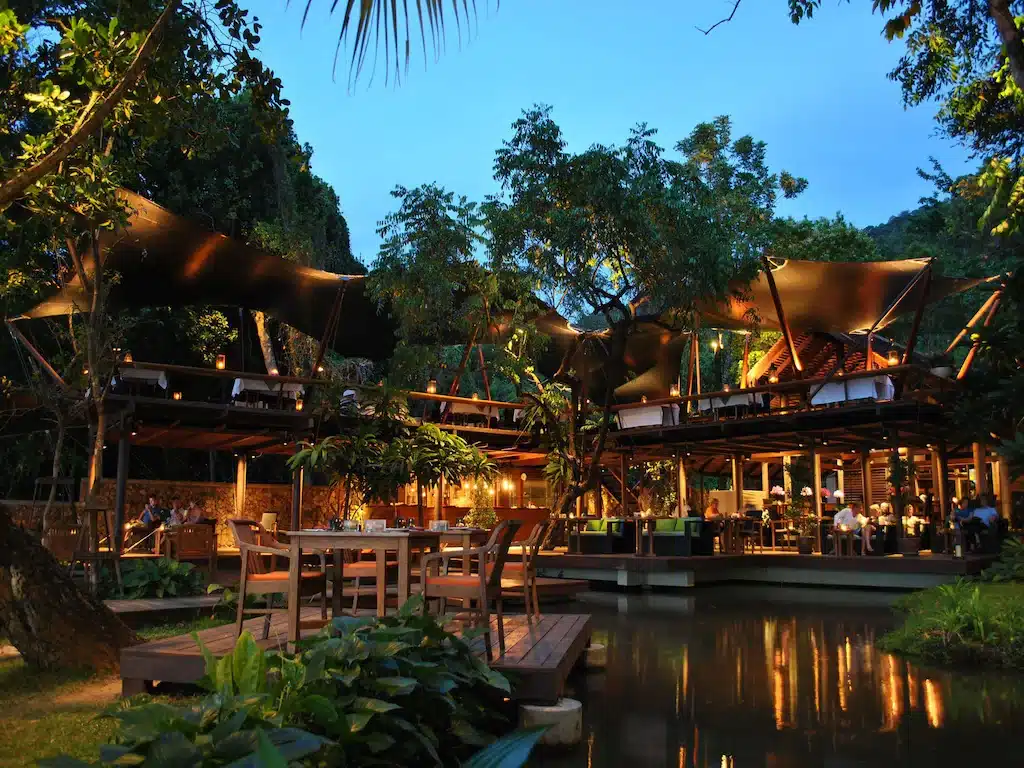 ร้านอาหารกลางแจ้งที่มีระเบียงไม้  ที่พักกระบี่ โต๊ะและเก้าอี้ ล้อมรอบไปด้วยต้นไม้เขียวขจีและบ่อน้ำ ส่องสว่างด้วยแสงไฟอันอบอุ่นในยามเย็น เหมาะสำหรับแขกที่มองหาประสบการณ์การรับประทานอาหารอันเงียบสงบที่ รีสอร์ทกระบี่.