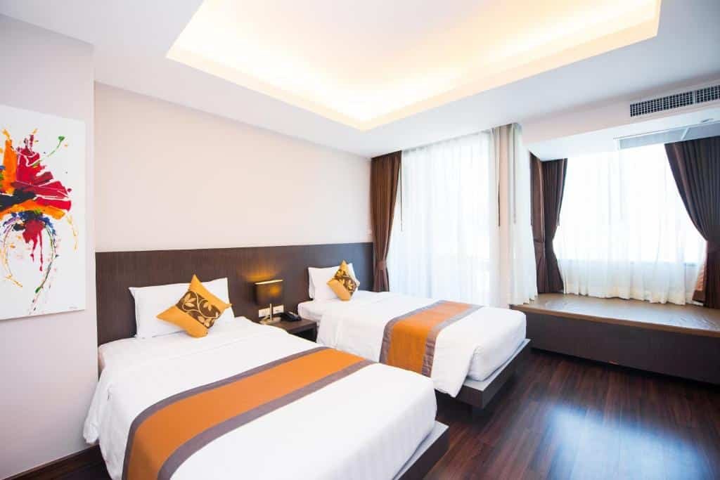 ห้องพักในโรงแรมทันสมัยที่โรงแรมกระบี่พร้อมเตียงเดี่ยว 2 เตียง ชุดเครื่องนอนสีขาวพร้อมรางสีส้ม โต๊ะหัวเตียง หน้าต่างบานใหญ่พร้อมผ้าม่าน และภาพวาดนามธรรมสีสันสดใสบนผนัง เหมาะสำหรับการพักผ่อนในกระบี่ครั้งต่อไปของคุณ.