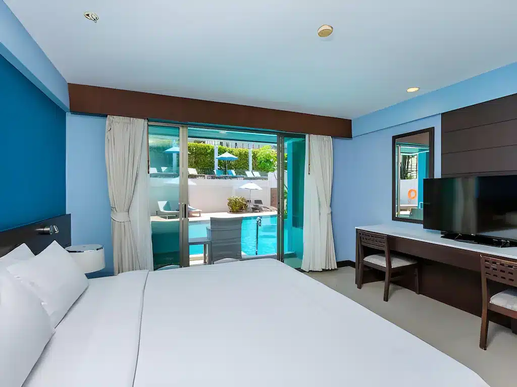 ห้องพักในโรงแรมที่มีเตียงนอนที่จัด ที่พักกระบี่ อย่างประณีต ทีวีจอแบนบนโต๊ะ และประตูกระจกที่เปิดออกไปสู่ลานพร้อมที่นั่งกลางแจ้งและวิวสระว่ายน้ำที่โรงแรมอันทันสมัยแห่งนี้.