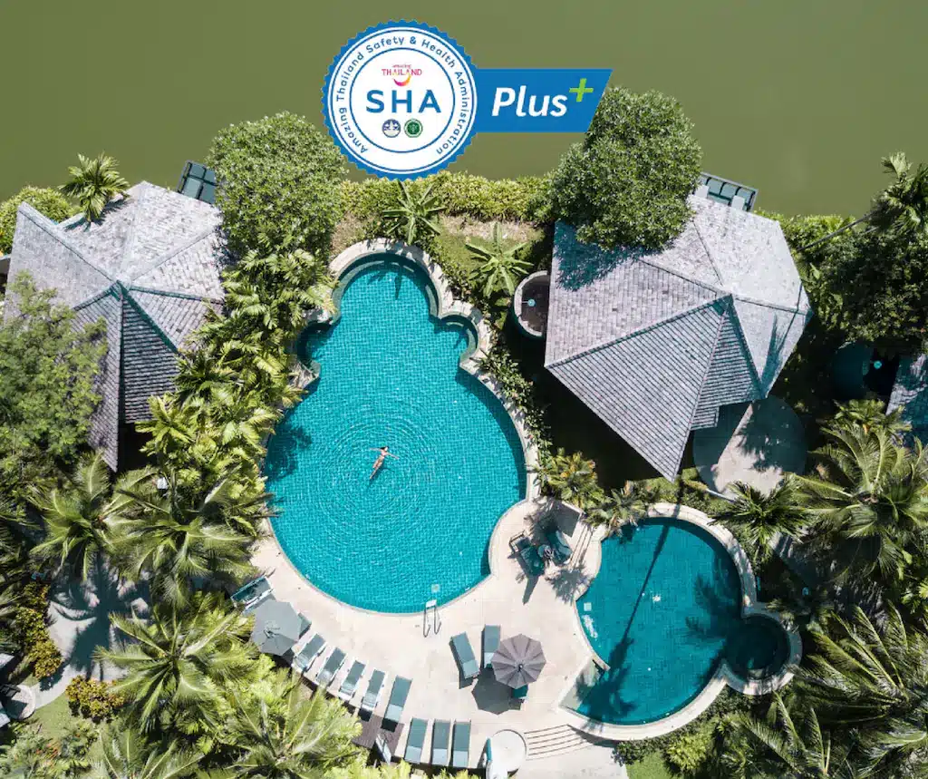 วิวทางอากาศของอ่าวนางสระว่ายน้ำของรีสอร์ทที่รายล้อมไปด้วยเก้าอี้นั่งเล่น  ที่พักอ่าวนาง ต้นปาล์ม และวิลล่า พร้อมด้วยคนว่ายน้ำ โลโก้การรับรอง SHA Plus+ ที่ด้านบน.