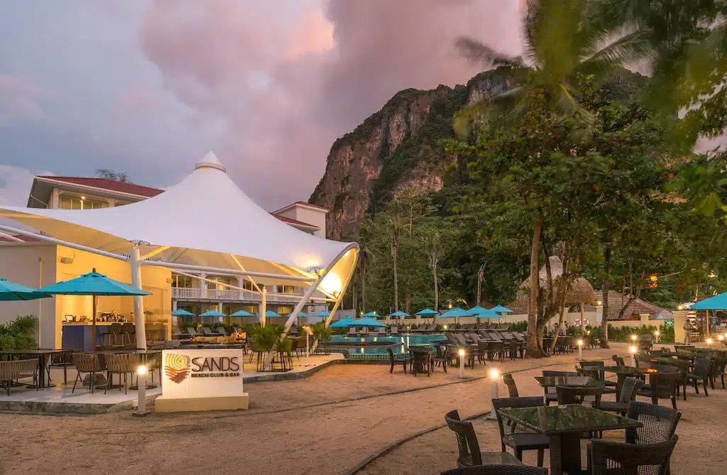 พื้นที่รับประทานอาหารกลางแจ้งที่ ที่พักอ่าวนาง ร้านอาหารริมชายหาดของแซนด์ส พร้อมด้วยโต๊ะ เก้าอี้ ร่มสีฟ้า และภูเขาที่สวยงามเป็นฉากหลังในยามพลบค่ำ เหมาะสำหรับแขกที่กำลังมองหาอ่าวนางกระบี่และประสบการณ์ริมทะเลที่น่าจดจำ.