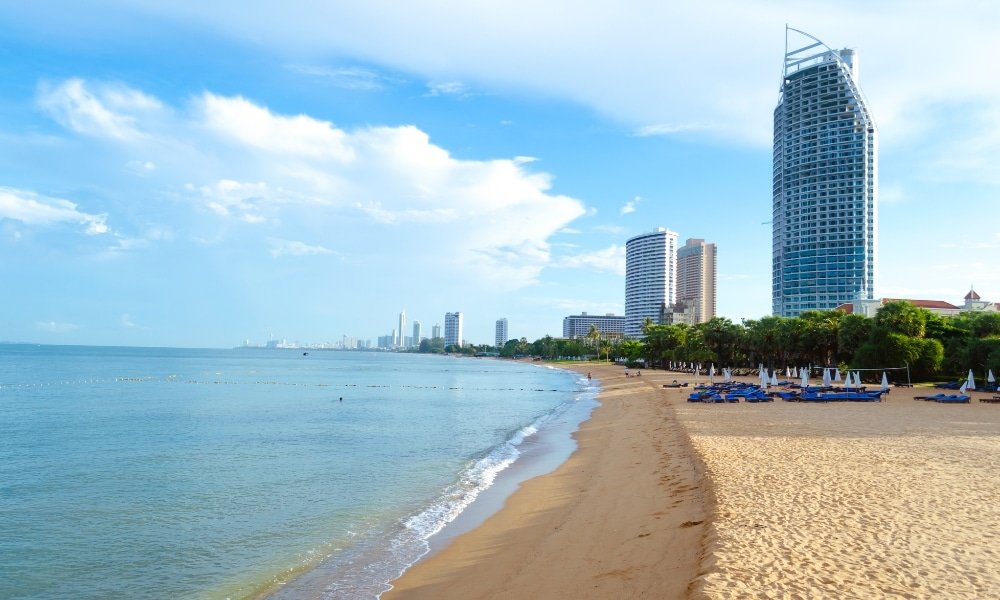 หาดทรายที่มีน้ำนิ่งสงบและมีตึกสูงระฟ้าเรียงรายอยู่ตามชายฝั่งภายใต้ท้องฟ้าที่มีเมฆบางส่วน เหมาะสำหรับผู้ที่มองหา ที่เที่ยวพัทยา ที่เที่ยวพัทยาติดทะเล.