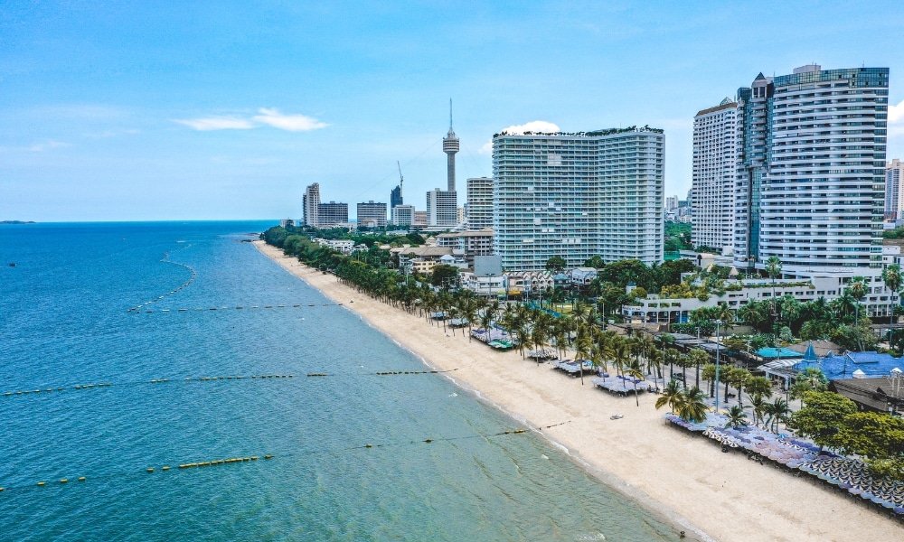 วิวทางอากาศของหาดทรายที่มีต้นปาล์ม น้ำทะเลสีฟ้าใส และตึกสูงระฟ้าตามแนวชายฝั่ง เหมาะสำหรับผู้ที่มองหา ที่เที่ยวพัทยาติดทะเล ในวันที่อากาศสดใส.