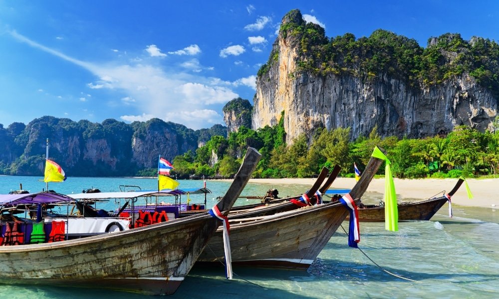 เรือหางยาวที่ทำจาก ที่พักอ่าวนาง ไม้สามลำพร้อมธงหลากสีสันจอดอยู่ในน้ำใสใกล้หาดทรายและหน้าผาหินปูน ซึ่งเป็นฉากที่โดดเด่นสำหรับนักเดินทางที่ค้นหาอ่าวนางหรืออ่าวกระบี่อ่าวนาง ท่ามกลางความงามอันเขียวชอุ่มของเขตร้อน.