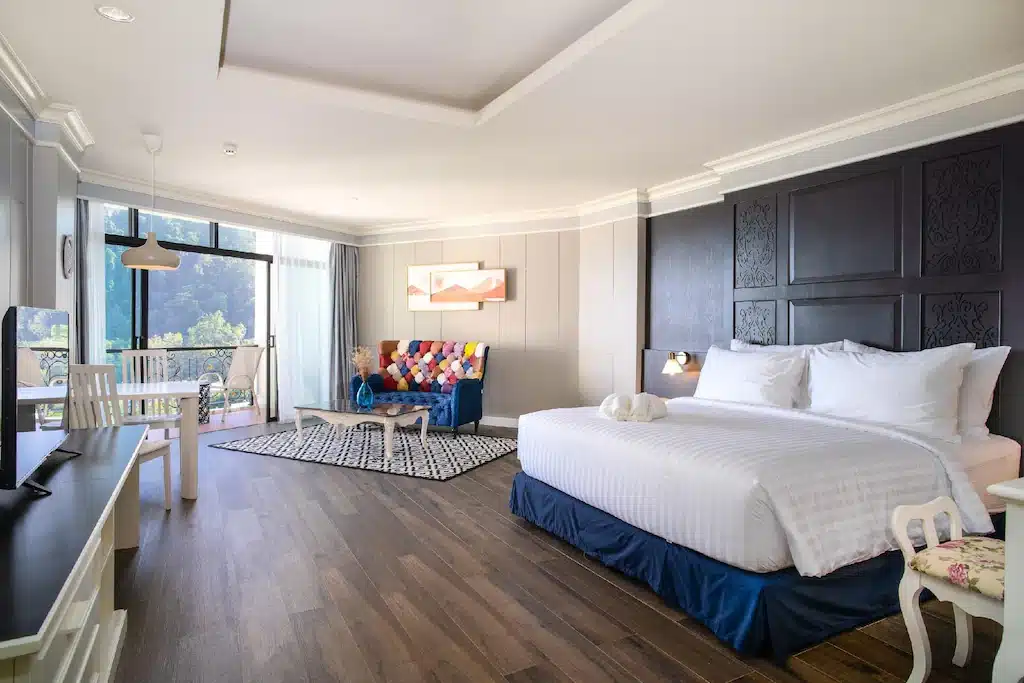 ห้องพักโรงแรมกว้างขวางของ โรงแรมกระบี่ มีเตียงขนาดใหญ่ พื้นที่รับประทานอาหารเล็ก โซฟาสีสันสดใส พื้นไม้ และหน้าต่างบานสูงจากพื้นจรดเพดานที่มองเห็นต้นไม้เขียวขจี.