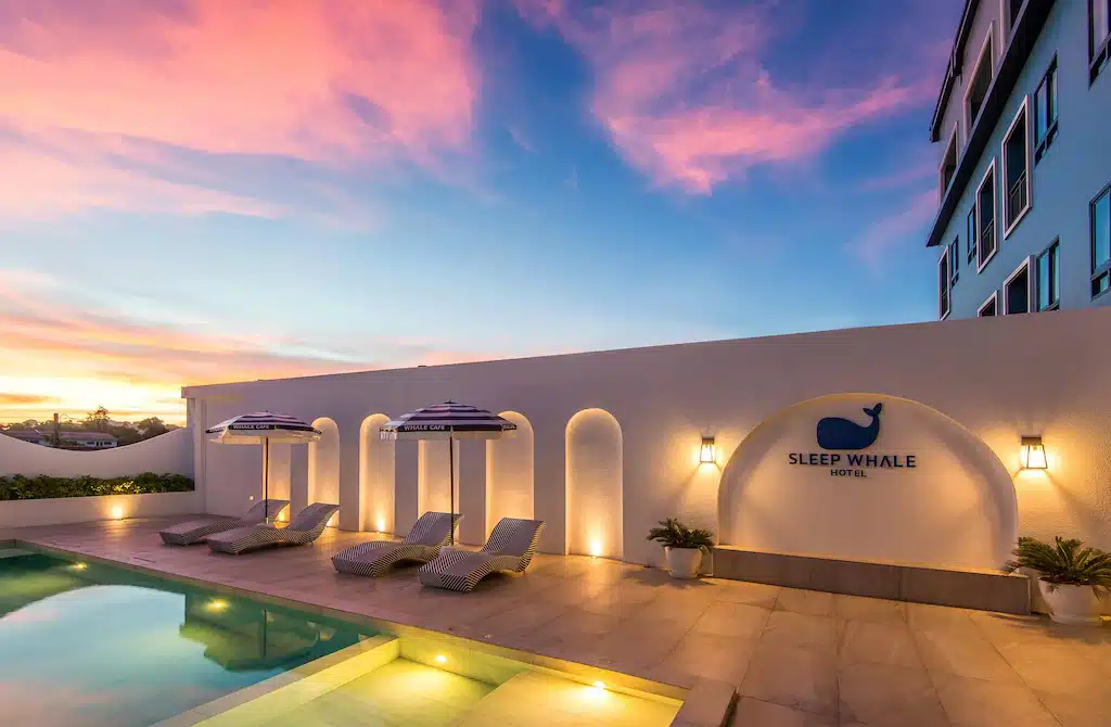 พื้นที่สระว่ายน้ำกลางแจ้งที่ Sleep Whale Hotel—รีสอร์ทกระบี่—พร้อม ที่พักกระบี่ เก้าอี้นั่งเล่น ร่ม ไฟติดผนัง และท้องฟ้าพระอาทิตย์ตกอันน่าทึ่งเป็นพื้นหลัง.