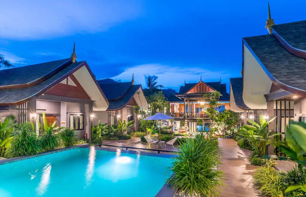 รีสอร์ทยามค่ำพร้อมอาคารสไตล์ไทยดั้งเดิม สระว่ายน้ำ ที่พักอ่าวนาง ที่มีไฟส่องสว่าง พื้นที่เขียวขจี เก้าอี้นั่งเล่น และศาลากลางที่มีแสงสว่างส่องสว่าง เหมาะสำหรับผู้ที่กำลังมองหาอ่าวนางราคาถูกหรือผ่อนคลายอ่าวนางกระบี่.