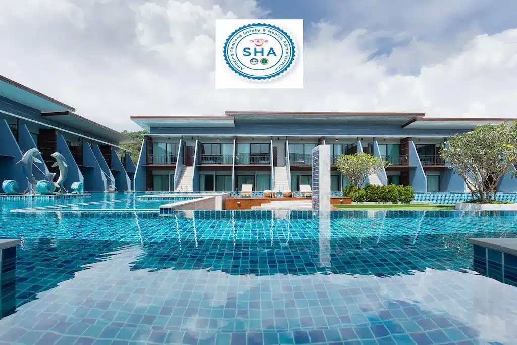 โรงแรมทันสมัยพร้อมสระว่ายน้ำกลางแจ้งขนาดใหญ่ น้ำทะเลสีฟ้าใส ที่นั่งริมสระ และโลโก้รับรอง SHA ที่ด้านบนตรงกลาง เหมาะสำหรับผู้ที่กำลังมองหา ที่พักกระบี่ อย่างมีสไตล์.