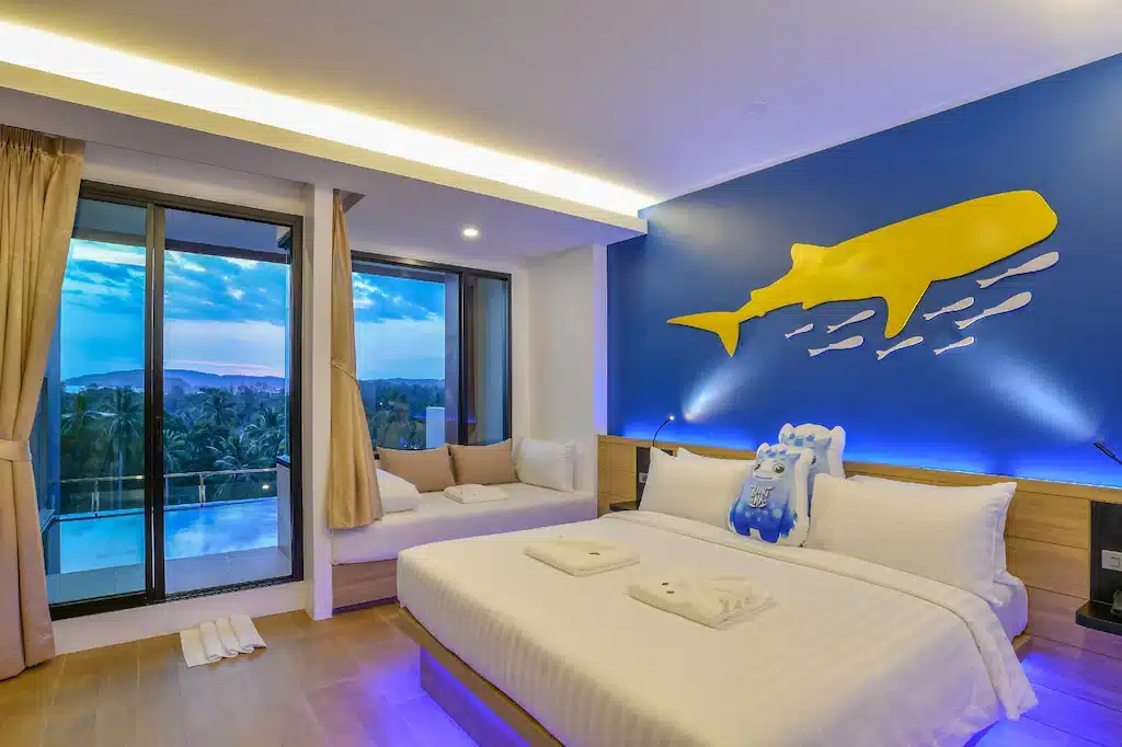 ห้องพัก ที่พักกระบี่ โรงแรมทันสมัยในโรงแรมเมืองกระบี่ มีเตียงขนาดใหญ่ โซฟา ประตูกระจกสู่ระเบียงที่มองเห็นทิวทัศน์ และผนังสีฟ้าตกแต่งด้วยลวดลายปลาวาฬและปลาสีเหลืองอันแสนสนุก.