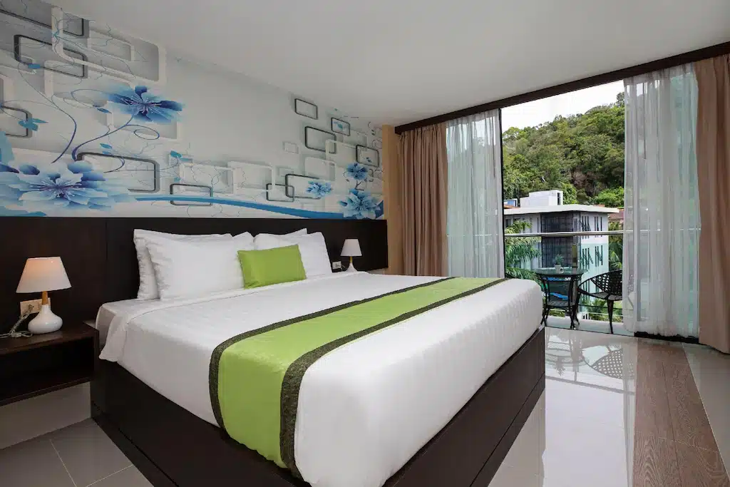 ห้องพั  โรงแรมกระบี่ กโรงแรมทันสมัยที่ โรงแรมกระบี่  มีเตียงขนาดใหญ่พร้อมชุดเครื่องนอนสีเขียวและสีขาว ผนังตกแต่งด้วยลายดอกไม้ โคมไฟข้างเตียง และระเบียงพร้อมที่นั่งกลางแจ้งที่มองเห็นต้นไม้เขียวขจี.