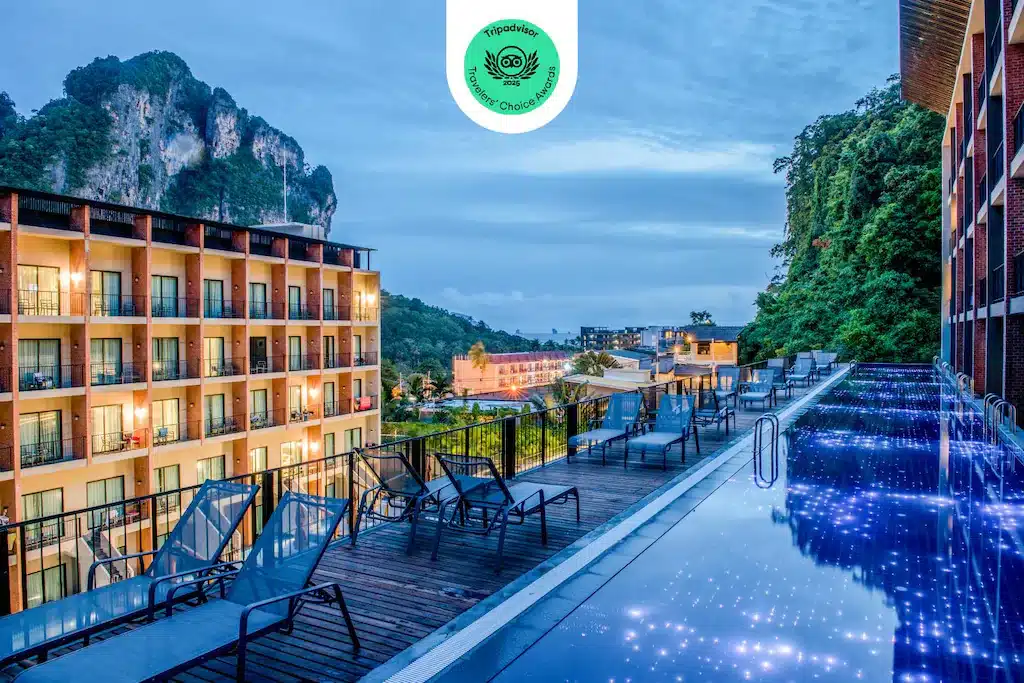 รีสอร์ท โรงแรมกระบี่ ทันสมัยกระบี่พร้อมระเบียงหลายระเบียงมองเห็นลานสระว่ายน้ำที่มีแสงสว่างพร้อมเก้าอี้เลานจ์ โดยมีฉากหลังเป็นเนินหินและแมกไม้เขียวขจีในยามพลบค่ำ.