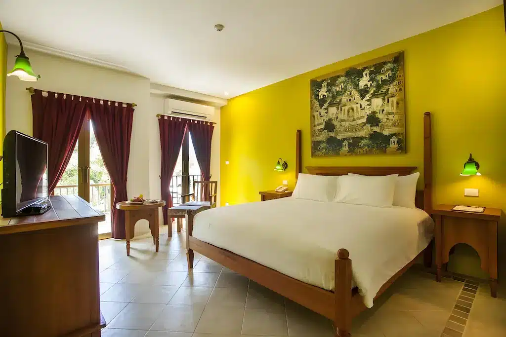 ห้องพักใน ที่พักอ่าวนาง โรงแรมสุดแสนสบายที่สเตย์อ่าวนาง มีเตียงคู่ ผนังสีเหลือง เฟอร์นิเจอร์ไม้ ทีวี งานศิลปะบนผนังอันสวยงามเหนือเตียง และหน้าต่างบานใหญ่ 2 บานพร้อมผ้าม่านสีแดง.