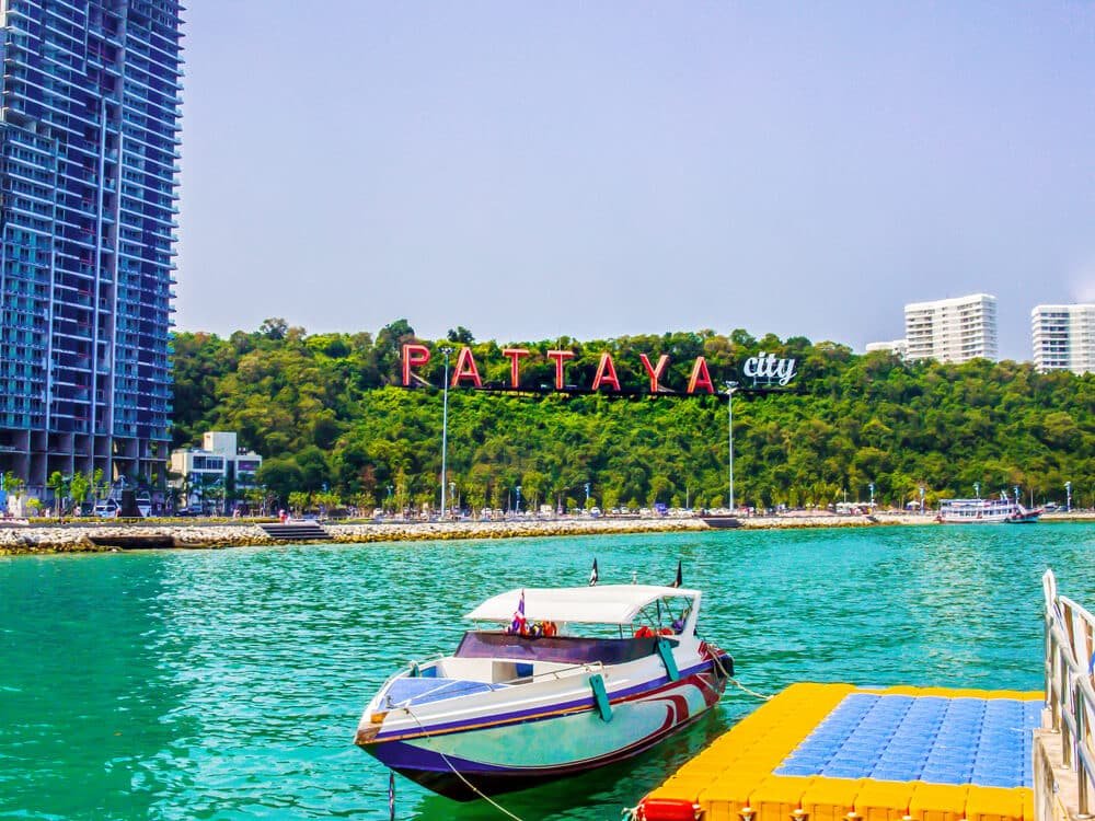 เรือเร็วจอดอยู่ในน้ำสีฟ้าใสใกล้กับแพลตฟอร์มลอยน้ำ พร้อมป้าย "เมืองพัทยา" อันเป็นสัญลักษณ์และอาคาร ที่เที่ยวพัทยา ทันสมัยเป็นฉากหลัง ซึ่งเป็นฉากที่สมบูรณ์แบบสำหรับใครก็ตามที่ต้องการสำรวจ ที่เที่ยวพัทยา.