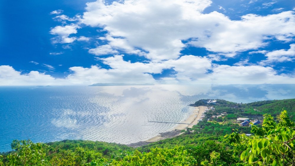 มุมมองทางอากาศของแนวชายฝั่งที่มีหาดทราย น้ำทะเลสีฟ้าใส และพืช เที่ยวพัทยา พรรณเขียวชอุ่มภายใต้ท้องฟ้าที่มีเมฆบางส่วน เหมาะสำหรับผู้ที่ต้องการสำรวจพัทยาที่เที่ยวและค้นพบความงามของที่เที่ยวพัทยา.