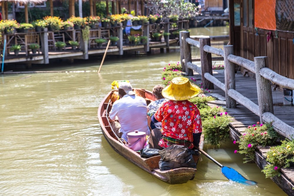 คนสามคนนั่งเรือไม้ล่องไปตามคลอง ที่เที่ยวพัทยา ข้างอาคารที่มีดอกไม้ คนที่พายเรือสวมหมวกสีเหลืองและเสื้อลายดอกไม้สีแดง ถ่ายทอดเสน่ห์ของ พัทยาที่เที่ยว ใกล้ทะเล.