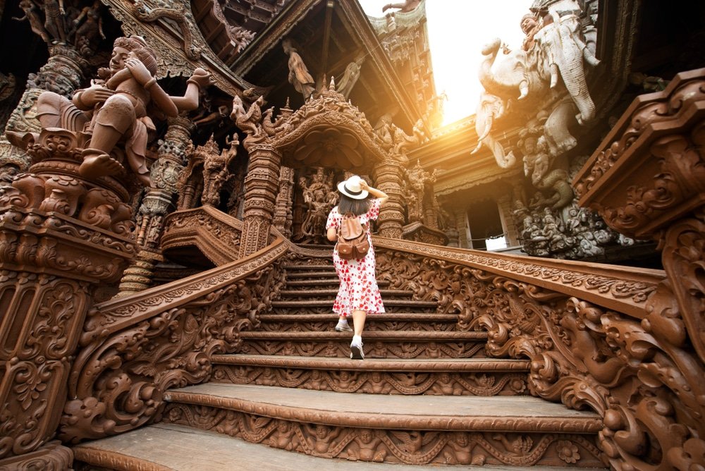 ผู้หญิงในชุดสีแดงและสีขาวเดินขึ้นบันไดไม้แกะสลักอย่างวิจิตรบรรจงใน ที่เที่ยวพัทยา วัด แสงแดดสาดส่องผ่านประติมากรรมที่มีรายละเอียด เป็นช่วงเวลาอันน่าทึ่งในการสำรวจ ที่เที่ยวพัทยา.