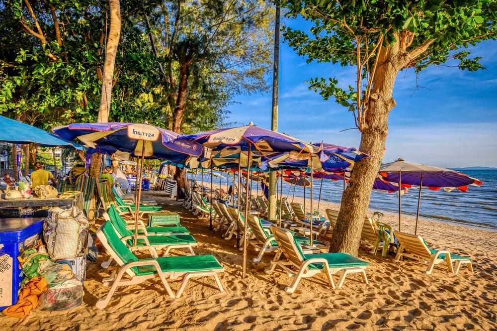 เก้าอี้เลานจ์สีเขียวและร่มสีน้ำเงินเรียงรายอยู่บนชายหาดทรายใกล้กับน้ำ ร่มรื่นด้วยต้นไม้ เป็นฉากที่น่าดึงดูดใจ เหมาะสำหรับใครก็ตามที่ต้องการมาท่อง เที่ยวพัทยา และผ่อนคลายริมทะเล.