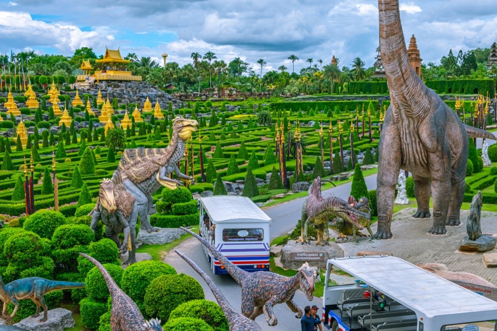 สวนสาธารณะแห่งหนึ่งใน พัทยาที่เที่ยว มีรูปปั้นไดโนเสาร์ขนาดใหญ่ รั้วต้นไม้ที่ได้รับการดูแลอย่างดี และรถรางสีขาวที่ให้นักท่องเที่ยวขับรถไปตามทางปูคอนกรีตภายใต้ท้องฟ้าที่มีเมฆบางส่วน.