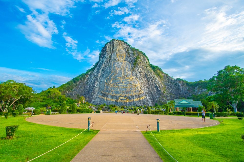 พระพุทธรูปสีทององค์ใหญ่แกะสลักบนภูเขาหินปูน รายล้อมไปด้วยต้นไม้และสนามหญ้าที่ได้รับการดูแลอย่าง เที่ยวพัทยา ดีภายใต้ท้องฟ้าสีฟ้าและเมฆหมอก เป็นจุดแวะพักที่สำคัญสำหรับผู้ที่ต้องการสำรวจ พัทยาที่เที่ยว และสถานที่ท่องเที่ยวที่เป็นเอกลักษณ์.