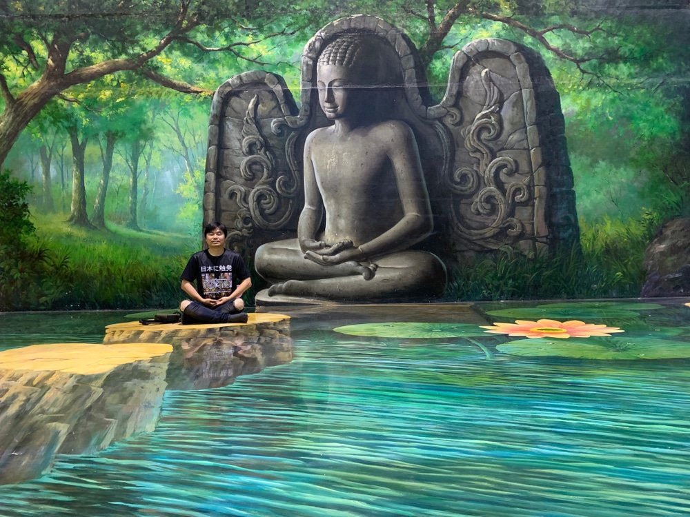 บุคคลหนึ่งนั่งขัดสมาธิบนหินขั้น ที่เที่ยวพัทยา บันไดทาสีหน้าจิตรกรรมฝาผนังขนาดใหญ่ของพระพุทธนั่งสมาธิ โดยมีสระน้ำและต้นไม้เป็นฉากหลัง ซึ่งเป็นฉากที่สร้างแรงบันดาลใจ ณ หนึ่งในที่เที่ยวพัทยาสำหรับสายชิลล์แห่งหนึ่ง.