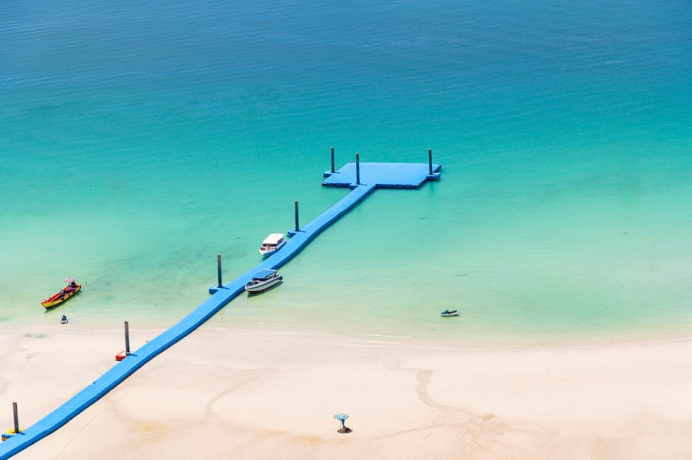 ท่าเรือสีฟ้ายาวทอดตัวเหนือน้ำทะเลสีฟ้าใสจากชายหาดทรายใน พัทยาที่เที่ยว มีเรือไม่กี่ลำจอดอยู่ใกล้ๆ และมองเห็นคลื่นเบาๆ.
