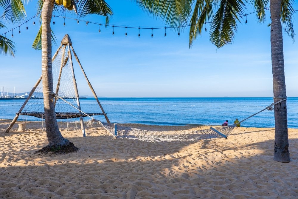 เปลญวนถูกแขวนไว้ระหว่างต้นปาล์มสองต้นบนชายหาดที่มีทรายขาว ที่เที่ยวพัทยา ละเอียด โดยมีน้ำทะเลสีฟ้าอันสงบและท้องฟ้าใสเป็นฉากหลัง เหมาะสำหรับการผ่อนคลายขณะเที่ยวชม ที่เที่ยวพัทยา.