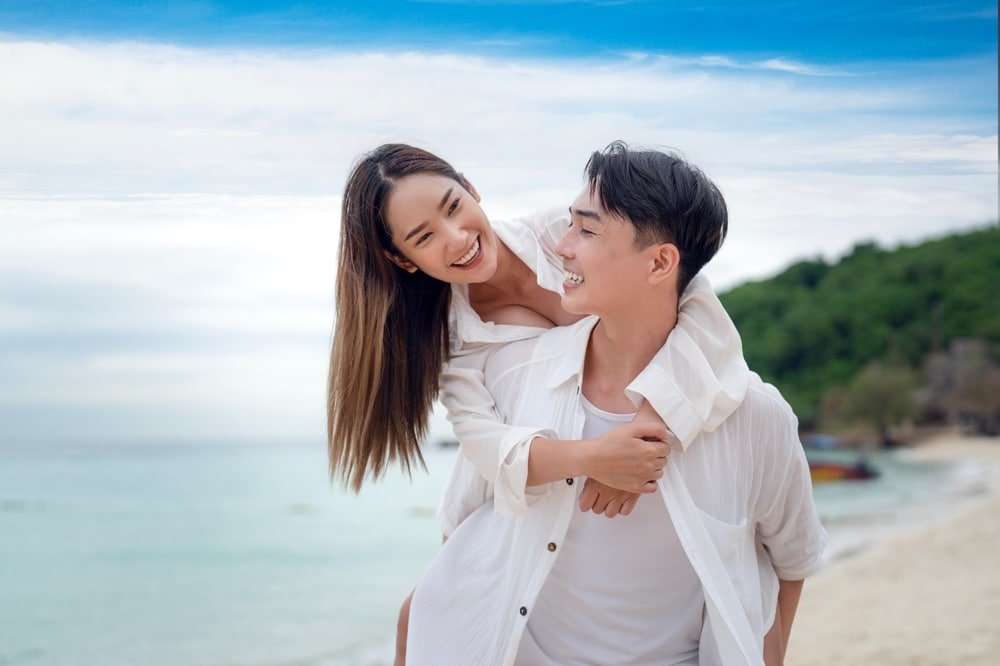 ผู้หญิงยิ้มขณะขี่หลังผู้ชายที่ชายหาด ทั้งคู่สวมเสื้อเชิ้ตสีขาว มหาสมุทรและความเขียวขจีเป็นฉากหลังที่สมบูรณ์แบบ รวบรวมความสุขของการเที่ยวพัทยาติด ที่เที่ยวพัทยา ทะเล.