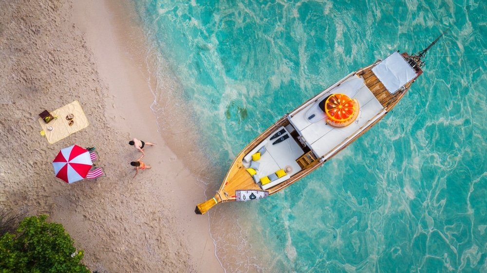 วิวทางอากาศของเรือจอดทอดสมอใกล้ เที่ยวพัทยา หาดทรายที่เที่ยวพัทยาติดทะเล พร้อมคนสองคน อุปกรณ์ปิกนิก และร่มสีแดงขาวข้างน้ำทะเลสีฟ้าใส.