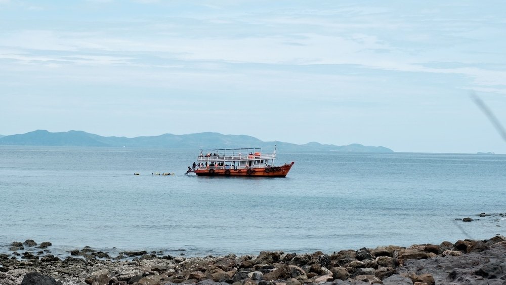 เรือสีแดงขาวลอยอยู่ในน้ำทะเลอันเงียบสงบใกล้แนวชายฝั่งหินพร้อม เที่ยวพัทยา เกาะต่าง ๆ ที่ห่างไกลมองเห็นได้ภายใต้ท้องฟ้าที่มีเมฆมาก จับภาพเสน่ห์อันเงียบสงบของที่เที่ยวพัทยา.