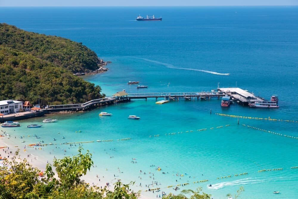 วิวชายหาดน้ำทะเลสีฟ้าใส ผู้คนเล่นน้ำ เรือใกล้ท่าเรือ และเรือใหญ่อยู่ไกลๆ ล้อมรอบด้วยเนินเขาเขียวขจี เหมาะสำหรับผู้ที่มองหา ที่เที่ยวพัทยาติดทะเล