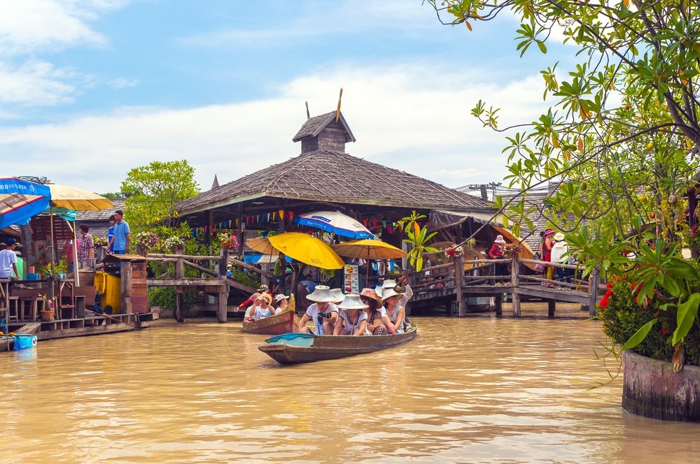 เรือไม้พร้อมผู้โดยสารสวมหมวกลอยอยู่ในแม่น้ำสีน้ำตาลใกล้ตลาดริมน้ำ ที่เที่ยวพัทยา ท้องฟ้าที่มีเมฆบางส่วน เป็นฉากอันสวยงามของ พัทยาที่เที่ยว ที่รอให้สำรวจ.