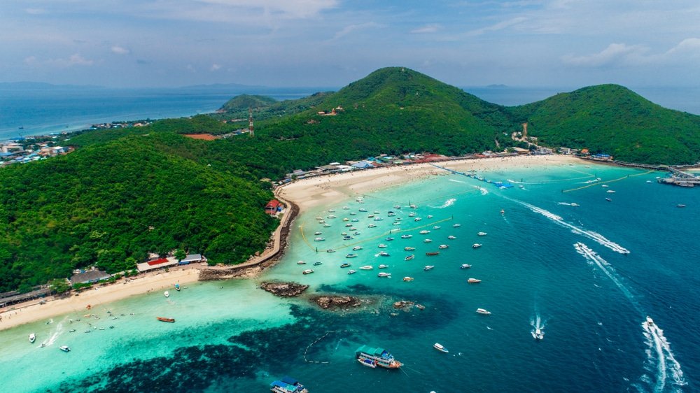 วิวทางอากาศของ เที่ยวพัทยา เกาะเขตร้อนใกล้ที่เที่ยวพัทยาติดทะเล มีเนินเขาเขียวขจี หาดทราย น้ำทะเลสีฟ้าคราม และเรือที่กระจัดกระจายอยู่ใกล้ชายฝั่ง.