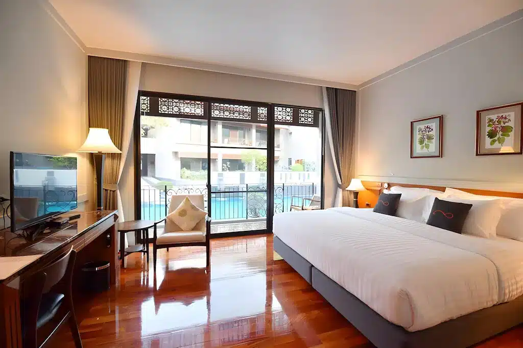 ห้องพักโรงแรมที่มีเตียงขนาดใหญ่,  ที่พักกาญจนบุรี โต๊ะทำงาน, โทรทัศน์, เก้าอี้นวม, และโคมไฟ, พร้อมประตูบานเลื่อนกระจกที่เปิดออกสู่ทัศนียภาพของสระว่ายน้ำ—เหมาะสำหรับการผ่อนคลายที่รีสอร์ทกาญจนบุรี หลังจากสำรวจบริเวณโดยรอบ.