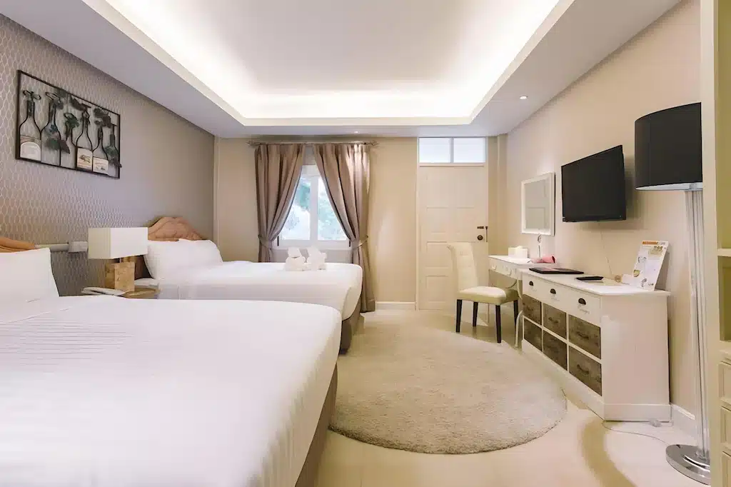 ห้องพักโรงแรมที่มีเตียงคู่สองเตียง, ทีวีติดผนัง, โต๊ะพร้อมเก้าอี้, พรมกลม, และหน้าต่างพร้อมผ้าม่านที่เปิดให้แสงธรรมชาติส่องเข้ามา—เหมาะสำหรับการพักผ่อนระหว่างท่องเที่ยว ที่เที่ยวใกล้กรุงเทพ สตูลหรือสำรวจสถานที่ท่องเที่ยวในจังหวัดสตูล.