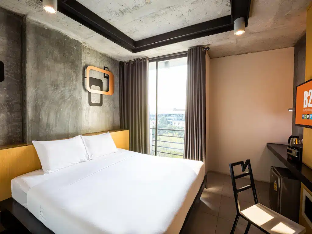 ห้องพักในโรงแรมทันสมัยพร้อมเตียงคู่ ผนังคอนกรีตสีเทา หน้าต่างพร้อมผ้าม่าน โต๊ะทำงาน เก้าอี้ และตู้เย็นขนาดเล็ก เหมาะสำหรับทุกคนที่กำลังมองหาที่พักพัทยาราคาถูกในบรรยากาศสบายและมีสไตล์.