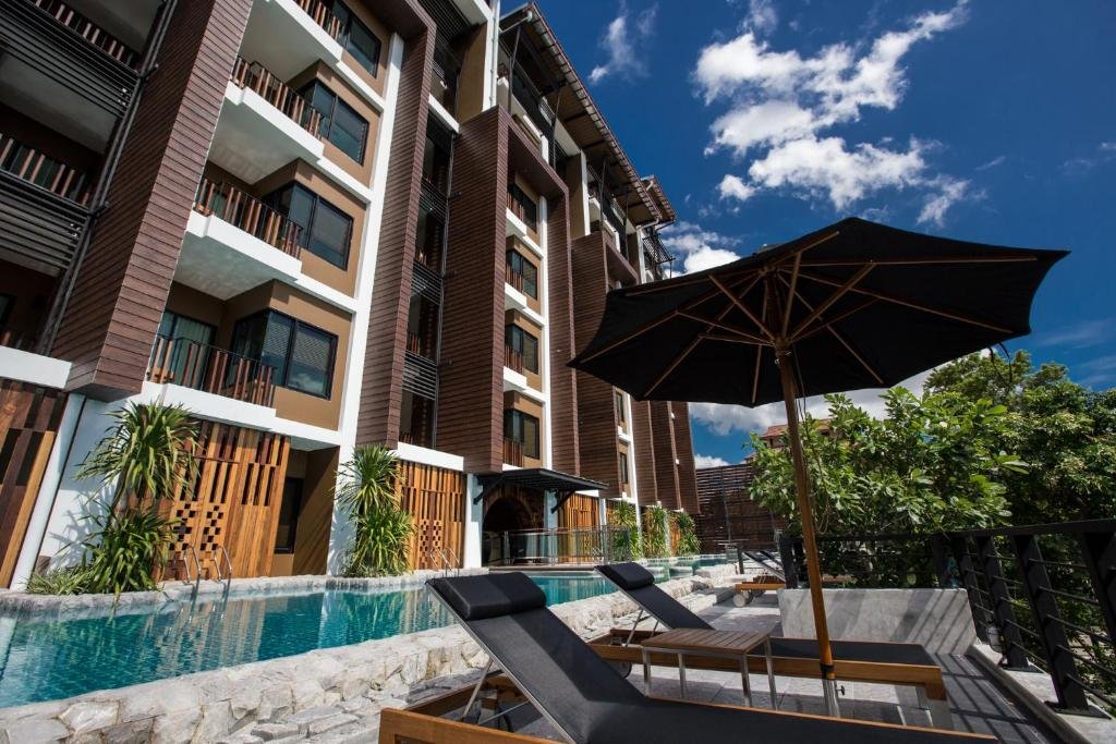 อาคารอพาร์ตเมนต์สมัยใหม่พร้อม ที่พักกาญจนบุรี ระเบียง เก้าอี้เลานจ์ริมสระว่ายน้ำ และร่มสีดำขนาดใหญ่ใต้ท้องฟ้าสีฟ้า—เหมาะสำหรับผู้ที่มองหาที่พักริมแม่น้ำกาญจนบุรีในบรรยากาศที่มีสไตล์และผ่อนคลาย.